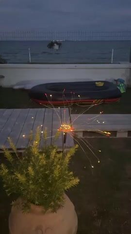 The video shows a dusk scene outdoors. In the foreground, a potted plant with green foliage is partially visible. A decorative light fixture resembling a dandelion with LED lights is positioned next to the plant. The lights on the fixture are illuminated. Behind the plant and light fixture, there is a wooden deck or pathway. Further back, there is an inflatable boat, black with red accents. Beyond the boat is a body of water, likely the sea, with a faint outline of a boat visible on the water. A fence runs along the edge of the water. The sky is a deep blue, indicating twilight. The overall atmosphere is calm and serene.