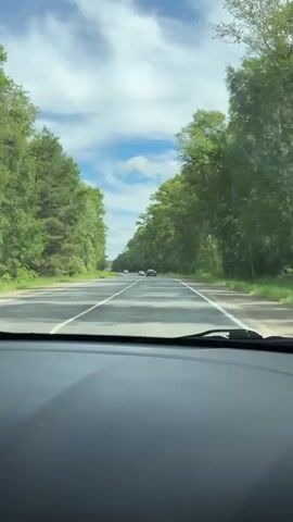 The video shows a first-person view from a car driving on a paved road. The road is bordered on both sides by dense green trees. The sky is partly cloudy with blue patches. Several cars are visible on the road ahead, also traveling in the same direction. The dashboard of the car is visible in the foreground. The time stamp indicates the video starts at 00:00 and ends at 00:04.
