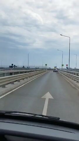 The video shows a first-person perspective from inside a vehicle driving on a bridge. The road ahead is empty except for a few cars in the distance. There are guardrails on both sides of the road, and streetlights are visible. There are also several road signs, including pedestrian crossing signs and warning signs. The sky is cloudy.