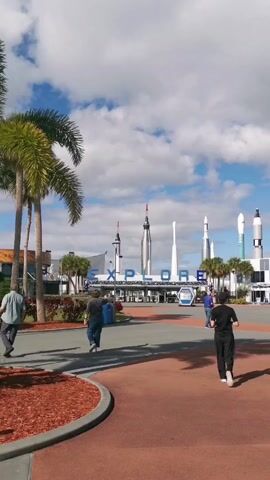 The video shows a plaza, likely at the entrance to a space center, under a partly cloudy sky. Several tall model rockets stand in the background behind the word "EXPLORE" in large blue letters. Palm trees are visible on the left side of the frame. People are walking around the plaza, some moving towards the entrance. A large screen displays an image on the right side of the frame. The plaza is paved with red bricks and gray concrete.