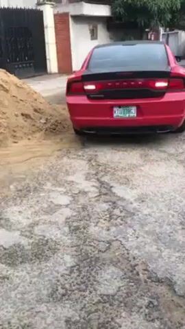 The video shows the rear of a red Dodge Charger parked on a cracked concrete surface. A pile of sand sits to the left of the car. A house with a black gate is visible in the background, and another vehicle is parked to the right. The car's license plate reads "KTU 778 GJ." The brake lights are activated. The scene appears to be a residential driveway or parking area.