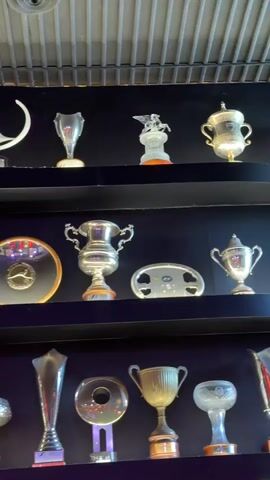 The video shows a wall with multiple shelves displaying a collection of trophies. The trophies vary in size, shape, and material, with many appearing to be made of polished metal. Some trophies have intricate designs, while others are simpler cup-shaped awards. The shelves are dark, creating a contrast with the gleaming trophies. The background above the top shelf shows a corrugated ceiling. There are no people visible in the video, and no actions or interactions are occurring.