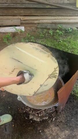 A large, silver pot sits over an open fire, filled with a stew containing meat and carrots. Steam rises from the pot, indicating it is cooking. The pot is placed on a metal stand, and the fire is built with logs and wood. In the background, a wooden fence is visible, and the ground is covered with dirt and grass. A black tarp is positioned behind the pot, possibly to shield it from wind or rain.