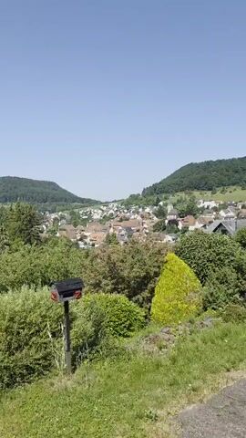 This media file presents a picturesque, elevated view of a small German village nestled amidst rolling green hills and lush vegetation. The scene is bathed in bright sunlight, suggesting a clear, warm day, likely mid-morning or early afternoon. A vast, clear blue sky dominates the upper portion of the frame, with only a few wispy clouds adding texture to the expanse.

In the foreground, a black mailbox with a red lid stands prominently on a wooden post, surrounded by dense, well-maintained green bushes and manicured hedges. Beyond this immediate area, the landscape unfolds to reveal the village. A cluster of buildings, characterized by their red-tiled roofs, is visible, indicating a traditional European architectural style. Interspersed with the houses are numerous trees, including tall evergreens and deciduous varieties, contributing to the verdant character of the settlement. Further in the distance, the scene is framed by the gentle slopes of tree-covered hills, suggesting a rural or semi-rural setting.

The overall impression is one of tranquility and natural beauty. The absence of people or overt activity implies a peaceful moment, perhaps a scenic overlook enjoyed by an unseen observer. The location cues, combined with the architectural style and the presence of the mailbox, strongly suggest a European, specifically German, setting, consistent with Albstadt, Germany. There is no visible text within the media file.