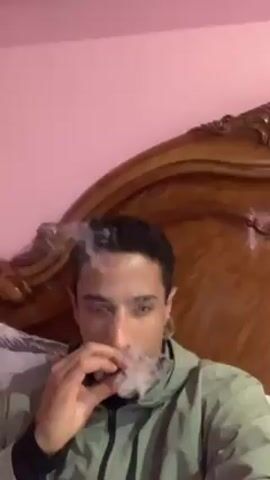 Here's a description of the video:
A man is lying in bed, smoking a cigarette. He is wearing a grey hoodie. The bed has a large wooden headboard. The walls behind him are painted pink. He exhales smoke and looks at the camera with a slight smile.