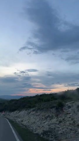 The video depicts a view from inside a vehicle, traveling on a paved road through a mountainous landscape. The road curves gently, and signage indicates sharp turns. The surrounding terrain consists of rocky slopes and dense green vegetation. In the distance, there are layered hills and mountains. The sky is predominantly blue-gray with scattered clouds, and the sun is setting. The overall environment appears serene and remote.