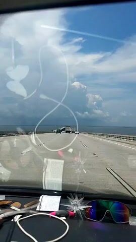 The view from inside a car driving along a bridge is captured. The road is straight, and a few other vehicles are visible ahead. The sky is partly cloudy, with the ocean visible on both sides of the bridge. A document, sunglasses, and a small decorative item are on the dashboard.
