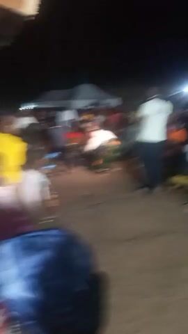 A group of people are gathered outdoors at night, illuminated by artificial lights and a tent. A few individuals are dancing in the center of the gathering. Some people are seated, while others stand watching. The ground appears to be dirt or gravel.
