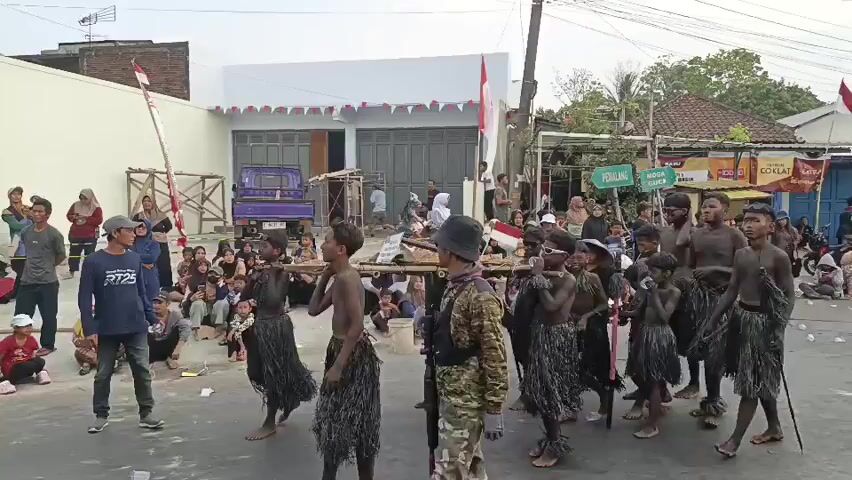 A group of young people, some appearing to be children, are participating in a procession or parade. They are dressed in dark, fringed skirts and have dark paint or mud on their skin. Several individuals are carrying a long wooden structure on their shoulders, which appears to be a makeshift stretcher or platform. Others are holding spears or long sticks. They are walking in formation down a street lined with spectators. The spectators are seated and standing, some of whom are holding Indonesian flags. The buildings in the background are decorated with Indonesian flags and banners. A purple truck is parked on the side of the street. The overall atmosphere suggests a cultural event or celebration.