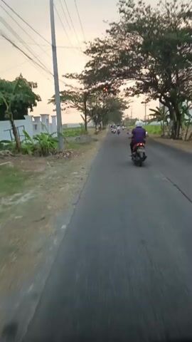 The video shows a first-person perspective of someone riding a motorcycle on a paved road. The sky is overcast with a warm, sunset-like glow. On the left side of the road, there are some short bushes and a white fence. On the right side, there are trees, some of which are bare and others with leaves, and houses. Several other motorcycles are visible in the distance on the road. The motorcycle being ridden is moving forward, and the surrounding environment is consistent with a rural or suburban road.