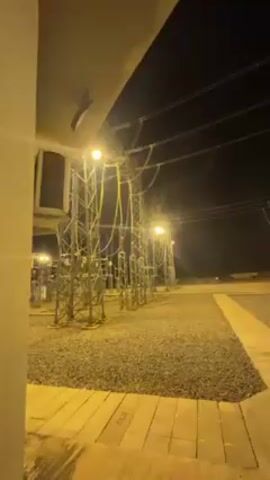 At night, the camera pans across a power substation.  The scene is illuminated by several bright yellow lights. The ground is covered in gray gravel. There are numerous metal structures with electrical components and wires. A concrete walkway extends into the scene.  The sky is dark and clear. There is no visible activity or people.