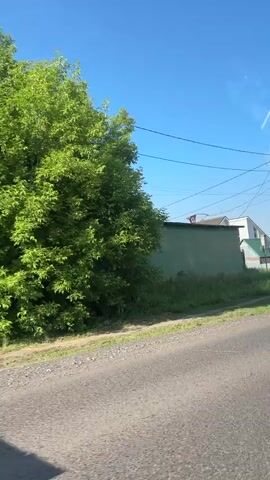 The video shows a street scene with a blue sky. On the right, a white car is driving on a road. In the background, there are houses with green roofs and a power pole with wires. A traffic light is visible on the left side of the image. There are trees on the left side of the road.