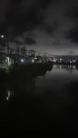 A nighttime landscape shot shows a body of water with a cityscape in the distance. The water is dark and reflects the lights of the city and the sky. The sky is filled with dark clouds that are illuminated by the city lights. The cityscape is composed of buildings and streetlights. There are also power lines and poles visible in the foreground. The overall atmosphere of the video is dark and mysterious.