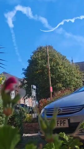 The video shows a clear blue sky with a few wispy clouds. A bright white contrail is visible in the sky, forming a curved shape. Below the sky, there is a large green tree with a tall, thin pole in front of it. To the left of the tree, there are buildings. In the foreground, the front of a silver car is visible. The car has a license plate that reads "HW17 FPF". There are also some plants in the foreground, out of focus.