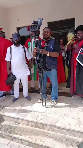 The video depicts a daytime gathering of people, likely associated with an academic event, at an outdoor, covered area in Unguwan Sanusi, Nigeria. A professional videographer, dressed in a blue vest, black t-shirt, and jeans, holds a video camera mounted on a tripod, occasionally smiling at the camera. He stands near a man in a white tunic, trousers, and sunglasses, who is holding a black bag.

Several individuals are prominently featured wearing academic regalia, including red gowns with green sashes and mortarboards. Another person is seen in a black academic gown with red and white trim. Other attendees are dressed in various formal and semi-formal attire, including traditional Nigerian garments.

The setting appears to be a modern building with large glass windows in the background, situated on a tiled patio or veranda with steps. People are seen conversing, mingling, and walking. The bright natural lighting suggests a clear, sunny day. Visible text on clothing and logos is present but not clearly legible.