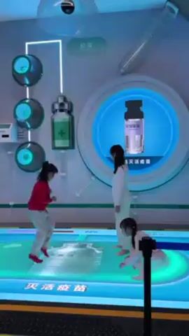 Three young girls play on a light-up platform in an interactive science exhibit. Two of the girls are small children, and the third appears to be a preteen. One child wears a red sweater and gray pants, another wears a white shirt and pink tutu, and the older girl wears white clothing. The platform displays different images at different times, including a vial of medicine, pink cells, and a cartoon nurse in a doctor's uniform. The surrounding walls feature additional science-themed exhibits, including round fixtures with illuminated green circles. A camera is visible on the ceiling. The overall lighting is bright and cool.