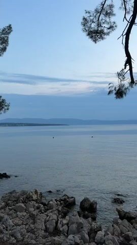 This video captures a serene waterscape at dusk, likely in Sveti Vid-Miholjice, Croatia. The scene is dominated by a calm, expansive body of water, possibly the Adriatic Sea, with a distant, hazy coastline of rolling hills or mountains. The sky is a soft gradient of blues and purples, with wispy clouds indicating the transition from day to night.

In the foreground, a rocky shoreline provides a textured contrast to the smooth water. A few scattered rocks are visible in the water itself, breaking the surface near the shore. Two figures are present in the water, appearing as dark silhouettes with their heads above the surface, suggesting they are swimming or wading. One figure, further to the left, is seen raising an arm, possibly waving or adjusting their position. The overall atmosphere is peaceful and tranquil, evoking a sense of calm and relaxation associated with a quiet evening by the sea. The time of day is clearly dusk or early evening, given the soft light and subdued colors. There is no visible text in the media file.