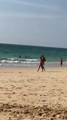 The video depicts a sandy beach under a bright, slightly hazy sky. The sea stretches out in the background, its blue-green water marked with white-capped waves. Several individuals are seen swimming or wading in the water. In the foreground, a couple walks along the shoreline. The woman appears to be wearing a bikini, while the man is wearing swim trunks. The overall scene suggests a casual, relaxing atmosphere on a sunny day at the beach.