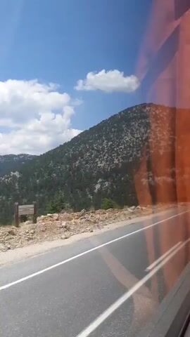 The video shows a view from a moving vehicle, likely a bus, looking out a window. The window has some reddish tinting, especially on the right side of the frame. The scene outside is a mountainous landscape with lush green trees covering the slopes. The sky is blue with scattered white clouds. A paved road with white lane markings is visible in the foreground, indicating the vehicle is traveling on a road that runs alongside the mountains. The terrain beside the road is rocky and uneven. The camera pans slightly to the left, revealing more of the rolling hills and mountains.