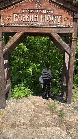 A woman walks down a stone pathway into a dense green forest through a rustic wooden archway. The archway has the words “АНГЕЛСКА ГРАДИНА” and “БОЖИЯ МОСТ” inscribed on it. The woman wears a striped top, dark pants, and carries a white bag on her shoulder. The day appears sunny, illuminating the bright green leaves of the surrounding trees and bushes. The camera remains stationary, capturing the woman as she disappears into the foliage.