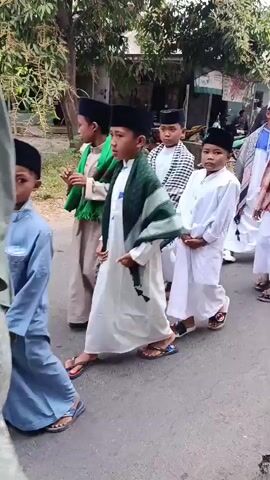 A group of young boys are walking down a paved road. They are all wearing white robes and black skullcaps. Some of the boys have scarves around their necks. The boy in the foreground on the left is wearing a green scarf. The boy in the middle is wearing a plaid scarf. The boy on the right is wearing a blue and white checkered scarf. There are trees and buildings in the background. The lighting suggests it is daytime.