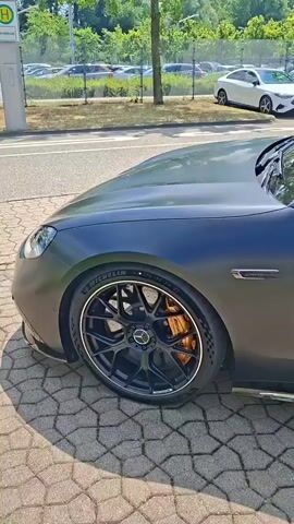 The video showcases the side profile of a matte black Mercedes-Benz coupe, featuring red interior seats visible through the tinted windows. The car is parked on a patterned brick driveway, with lush green grass bordering the area. In the background, a row of cars is visible in a parking lot, with trees and a bright blue sky overhead, suggesting a sunny day. A white Mercedes sedan is parked prominently in the background. The car's design highlights its sporty features, including custom black rims with gold brake calipers and a sleek side profile. A small emblem on the fender reads "V8 BITURBO 4MATIC+". The overall atmosphere is one of luxury and automotive appreciation, captured during daylight hours in what appears to be an outdoor setting, possibly a dealership or a private residence in Rastatt, Germany.