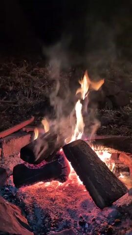 The video begins with a close-up shot of a campfire burning in the dark. Logs are arranged in a pyramid, and flames are visible, along with smoke rising from the fire. Then, the video shifts to show an exterior view of a window. The window has a white frame and appears to have a brick-patterned blind or covering. A speaker sits on the windowsill, and colorful lights are shining, creating rainbow patterns on the blinds. The wall surrounding the window is stucco. Music is playing.