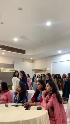 The video shows an indoor gathering of people, primarily women, seated at round tables covered with white tablecloths. In the foreground, several women are visible, some looking towards the front of the room. The setting appears to be a banquet hall or event space, with white walls, a tiled floor, and overhead lighting. In the background, a few women are standing and walking, and there are some decorative elements like golden stands and what appear to be beverage dispensers. The overall atmosphere seems casual and social, possibly a party or a celebration.