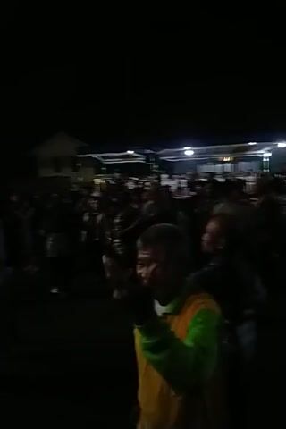Here is a matter-of-fact description of the video:

The video shows a nighttime scene, probably an outdoor concert or festival, judging by the large number of people visible in the crowd.  The scene is lit by a row of bright lights. The camera moves through the crowd, briefly focusing on individuals. People are mostly standing and interacting, with some appearing to be singing along with music that is playing in the background.