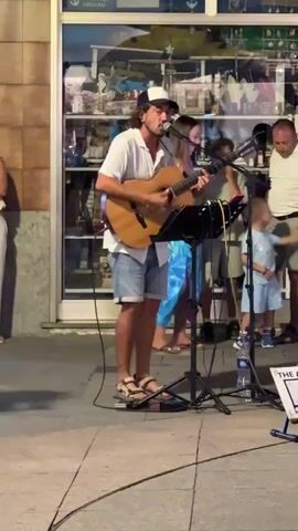 A man is playing an acoustic guitar and singing into a microphone on a sidewalk. He is wearing a white t-shirt, blue denim shorts, a baseball cap, and sandals. He is standing in front of a music stand with a microphone. Behind him is a storefront with a large window displaying various items. Several other people are visible in the background, including a man walking by on the left, and a child standing near the musician. The ground is paved with what appears to be cobblestones. The lighting suggests it is daytime or early evening.