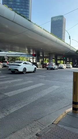 The video shows a street intersection in a city, underneath an elevated highway or bridge that has greenery on top. Traffic lights are visible. A black and white bus crosses the crosswalk and makes a left turn. Several cars are at the intersection, including a white sedan, a white SUV, and a silver minivan. The street is paved and has crosswalk markings. There are tall buildings in the background. The sky is blue and the weather appears sunny. A bollard is in the foreground of the shot.