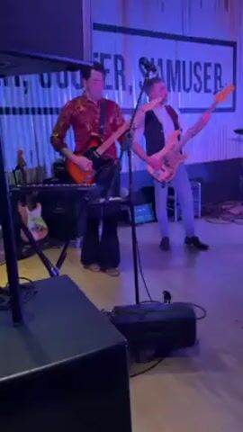 A medium shot captures a male band member playing an orange electric guitar with fervor, standing in front of a keyboard on a stage bathed in blue light. He wears a patterned reddish-gold shirt, dark pants, and tan shoes. A microphone stands before him, attached to a tall stand. A second band member is partially visible to the right, playing a bass guitar while wearing a vest and jeans. Behind the musicians is a wall with white writing visible, partially obscured by the bright light. A speaker and other equipment are placed to the left and bottom of the frame. The camera remains relatively stable throughout the recording.