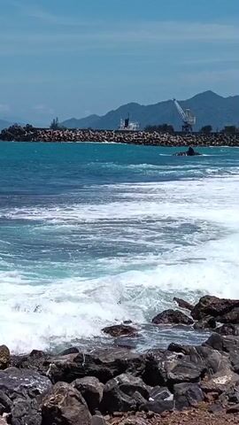 The video shows a coastal scene with waves crashing on a rocky shore. In the background, there is a breakwater made of large rocks, with ships and cranes visible on the other side, suggesting a harbor or port. Mountains rise behind the port. The sky is blue with some clouds, and the water is a vibrant turquoise. The camera is positioned low, looking out towards the sea and the breakwater.