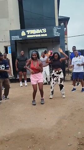 A group of approximately ten people are gathered outside a building with a sign that reads "TREDA'S SHOPPING HUB." Two women are in the foreground, dancing energetically. The woman on the left wears a red tank top and pink shorts, while the woman on the right wears a black crop top and patterned pants. Several other individuals, mostly men, are standing and watching, some appearing to dance as well. The environment is outdoors, with a dirt or gravel ground and a building facade behind the group. The overall atmosphere appears to be casual and celebratory, possibly a street performance or a flash mob.
