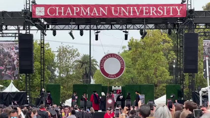 The scene shows a Chapman University graduation ceremony taking place outdoors on a stage. Above the stage hangs a red banner with the words "CHAPMAN UNIVERSITY" in white. A large, circular Chapman University seal is positioned behind the stage. Several individuals in academic gowns are standing on the stage. A crowd of graduates and attendees are facing the stage, many holding up their phones to take pictures or videos. Speakers are positioned on either side of the stage. The sky is overcast, and there are trees visible in the background.