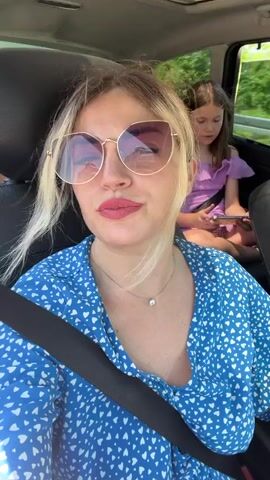 This is a close-up video of a woman with blonde hair and sunglasses, wearing a blue dress with a white heart pattern. She is sitting in the passenger seat of a car, with a seatbelt across her chest. Her hair is blowing in the wind, suggesting the car is moving. In the background, a child is visible in the back seat, holding a phone. The setting appears to be a road trip through a rural or natural landscape, with greenery and a body of water visible through the car window. The woman is speaking or singing, with an animated expression. The time of day appears to be daytime, and the weather seems pleasant. The emotional tone of the video is lively and energetic. No text is visible in the media file.