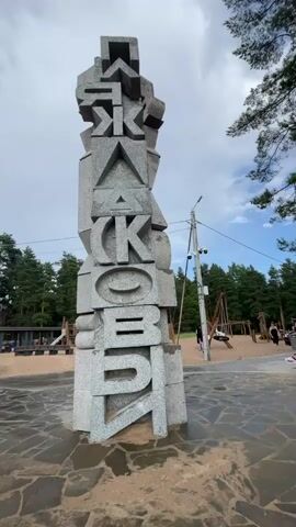 A tall, abstract sculpture made of grey stone blocks with carved letters stands in the foreground. The letters appear to be in the Cyrillic alphabet and are stacked vertically, forming a word. The sculpture is positioned on a stone-paved area.

In the background, there is a playground with wooden structures and sand. Several people are visible in the playground area, some walking and others sitting on benches. The scene is set outdoors with a backdrop of lush green trees, suggesting a park or recreational area. The sky is partly cloudy, indicating daytime. The overall atmosphere appears relaxed and casual, with people enjoying the outdoor space. The presence of a playground and people suggests a family-friendly environment. The sculpture itself is a prominent feature, likely a landmark or artistic installation within the location. The time of day is likely late morning or afternoon, given the lighting and the activities of the people. The location cues, such as the trees and the playground design, combined with the provided location context of Solnechnoye, Russia, help place the scene.