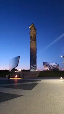 The video shows a war memorial at dusk. The memorial consists of a tall rectangular pillar topped with a statue, two curved mirrored walls, and an eternal flame. People are walking around the memorial plaza. The sky is a deep blue, with hints of orange on the horizon. Streetlights illuminate the plaza.