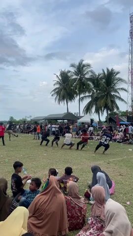 The video shows a tug-of-war competition taking place outdoors on a grassy field. A group of men, divided into two teams, are pulling a rope. One team is on the left side of the frame, dressed in dark clothing, while the other team, on the right, is wearing varied colored shirts, one with the number 83 on it. Both teams are exerting significant effort, with their bodies strained and faces showing exertion. In the background, a crowd of spectators, including many women and children wearing headscarves, are gathered to watch the event. Palm trees are visible behind the competitors, and tents are set up in the distance. The sky is overcast with some clouds. The overall atmosphere appears to be that of a community event or festival.
