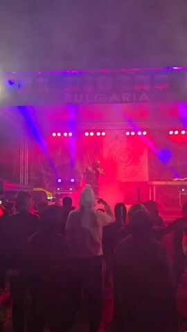 The video shows a singer performing on stage in front of an audience. The singer is wearing a black shirt, vest, and baseball cap, and has a microphone in hand. The stage is bathed in red and purple lights and smoke. Behind the singer is a backdrop with the words "KHYBOV SOUND" repeated. Audience members are visible in the foreground, some holding up phones to record the performance. The overall atmosphere is energetic and concert-like.