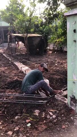 A person wearing a green t-shirt, grey pants, a dark baseball cap, and sandals is kneeling on the ground, working on a construction site. They are using a tool to place a white brick into a trench. Rebar is laid out in front of them on the ground. In the background, there is a partially built structure with rebar and concrete blocks, piles of dirt, trees, a house, and a light green painted concrete pillar. The sun appears to be shining.