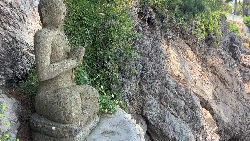 A video clip begins with a close-up of a stone statue of the Buddha in a meditative pose, situated amongst rocks and greenery. The camera then pans to the right, revealing a rocky coastline with lush vegetation and trees. The turquoise waters of the sea are visible, with a few people swimming. The camera continues to pan, showing more of the coastline and the open sea, with several rocks protruding from the water. The clip ends with a view of the sea and a distant coastline under a clear blue sky. A paddleboard is visible on the concrete surface in the foreground.