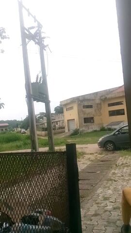 This is a slightly tilted, vertical video clip of an outdoor scene in Ire, Nigeria. The foreground is dominated by a large electrical pole with transformer equipment. Beyond the pole, there are several weathered, multi-story buildings constructed from concrete, with some parts painted yellow. The buildings have visible windows and a large roller-shutter door on one of them.

The scene appears to be in a semi-urban or industrial area. There is green grass and some trees visible, suggesting it's daytime. The sky is overcast, indicating cloudy weather. A dark metal mesh fence is also visible on the right side of the frame. In the background, a silver car is parked on a paved surface next to one of the buildings. A person's yellow-clad leg is visible at the very top right of the frame, suggesting they are standing or walking through the scene.

The overall impression is one of a functional, perhaps older, infrastructure setting in Nigeria. The video is mostly still, with slight camera movement. There is no visible text in the clip.