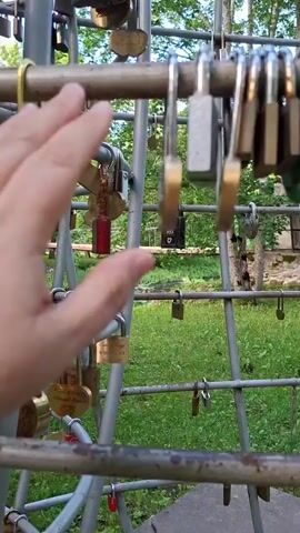 A hand is shown touching a green padlock on a metal structure covered in many padlocks. The structure appears to be outdoors with grass and trees in the background.