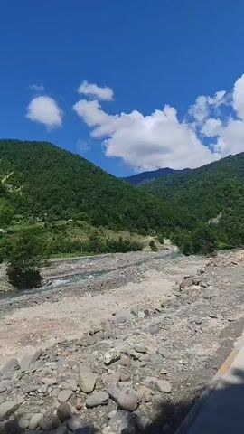 The video shows a wide, rocky riverbed with a shallow stream flowing through it. The river is surrounded by lush green mountains under a bright blue sky with scattered white clouds. The foreground is filled with various sized grey and brown rocks and pebbles. The water in the river appears to be a light blue or turquoise color. The overall scene depicts a natural, mountainous landscape on a sunny day.