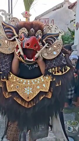 A person wearing an elaborate, colorful mask resembling a boar or wild pig, adorned with gold and red detailing and a headdress of palm fronds, stands outdoors. The mask has large tusks, wide eyes, and a gaping mouth. The person is also wearing a black shirt and a decorated collar that appears to be part of the costume. The background is slightly blurred, suggesting it's out of focus, but shows hints of buildings and other people, indicating a public space, possibly a street or courtyard during an event or festival. The lighting suggests it is daytime.