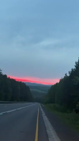 The video shows a scenic view of a road leading into a valley at dusk. The sky is a mix of gray clouds with a vibrant band of red near the horizon, indicating either a sunset or sunrise. Dense green trees line both sides of the asphalt road, which has a yellow line marking the center. In the distance, rolling hills are visible, also covered in greenery. The road appears to be empty, with no vehicles or people visible. The overall impression is one of a peaceful, natural landscape during twilight.