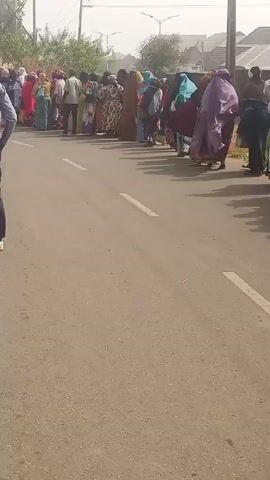A crowd of people, mostly women in colorful traditional clothing, are lined up on a paved road. The camera follows a man in a blue outfit with a backpack, walking towards the line. A water tower is visible in the background. The scene appears to be outdoors on a slightly hazy day.