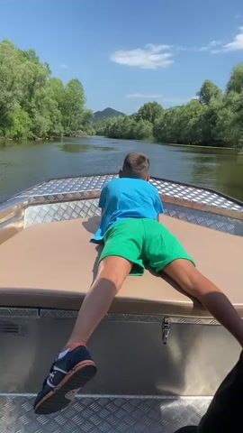 A boy wearing a blue t-shirt and green shorts lies on his stomach on the front of a boat. The boat is moving down a river with trees on either side. The sky is blue with some clouds. The boy is facing away from the camera.