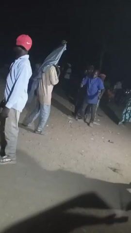 The video shows a group of people dancing in what appears to be a dirt-covered area at night. There are about eight individuals visible, most of whom are in motion. One man wears a red baseball cap, a light-colored button-down shirt, and gray pants. Another man wears a blue shirt and dark pants. A woman wears a white shirt and a colorful patterned skirt. They are surrounded by other people, some of whom are partially obscured by the darkness. The lighting is dim, making it difficult to see details. There is a single light source that appears to be coming from above, creating stark shadows on the ground. The people are moving and gesturing to music that is playing in the background.